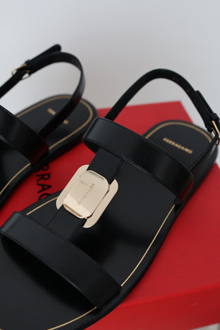 Ferragamo Capri black leather flat sandals in Size 8 featuring gold-tone Gancini hardware, open toes and adjustable slingback straps, displayed on the original red Ferragamo box.