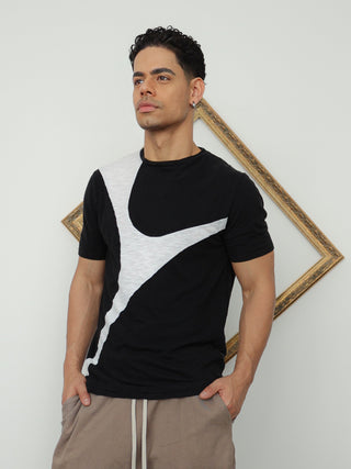 A man modeling a black and white color-blocked short-sleeve t-shirt paired with taupe cropped shorts, black socks, and black sneakers, standing against a white wall with a gold geometric frame.