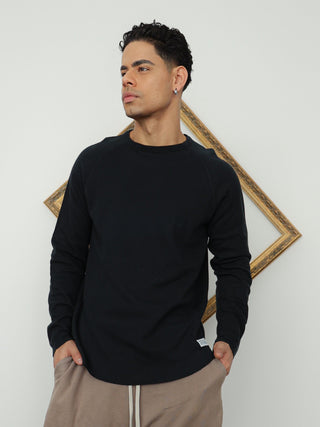 A man modeling a black long-sleeve crewneck sweater, taupe drawstring shorts, gray compression leggings, and black sneakers, standing against a white wall with a gold geometric frame.