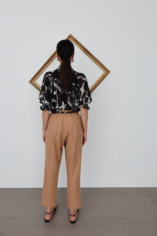 Model wearing Acne Studios camel tapered wool trousers with front pleats, styled with a black printed blouse; close-up images show pocket detailing and waistband.