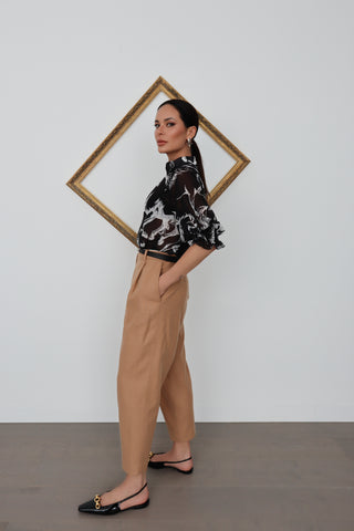 Model wearing Acne Studios camel tapered wool trousers with front pleats, styled with a black printed blouse; close-up images show pocket detailing and waistband.