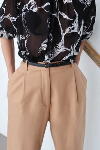 Model wearing Acne Studios camel tapered wool trousers with front pleats, styled with a black printed blouse; close-up images show pocket detailing and waistband.