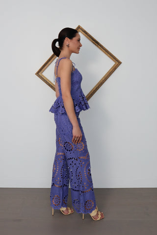 Model wearing Zimmermann violet embroidered linen set with peplum top and flared trousers, standing against white wall with gold tilted frame