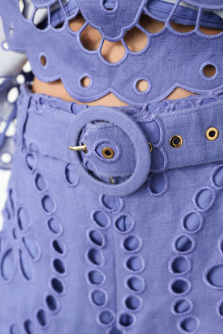 Model wearing Zimmermann violet embroidered linen set with peplum top and flared trousers, standing against white wall with gold tilted frame