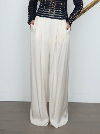 A woman stands in a minimalist room wearing a dark navy long-sleeve top with gold metallic chain detailing across the chest and wide-leg cream trousers. She is positioned in front of a white wall with a tilted gold picture frame behind her. Shona Joy Satin Pants.