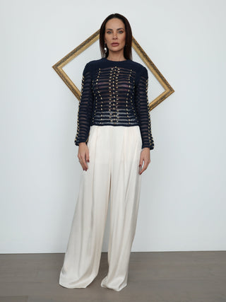 A woman stands in a studio wearing a dark navy long-sleeve knit top with horizontal metallic gold chain details across the chest and sleeves, paired with high-waisted cream wide-leg trousers. She is posed in front of a white wall with a gold-framed geometric art piece behind her.
