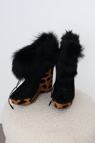 A pair of black fur-trimmed  Prada ankle boots with leopard-print uppers, displayed on a round white textured ottoman against a white wall.